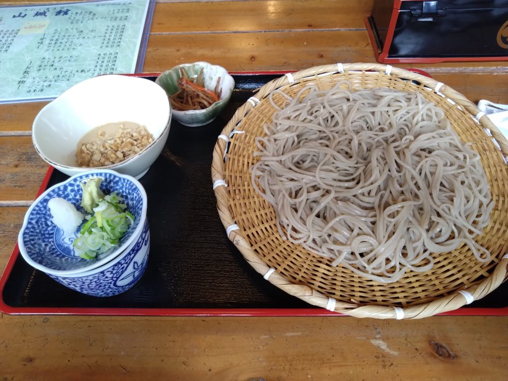 ざるに盛られたクルミ蕎麦と薬味、小鉢が木のテーブルの上に並んでいる。